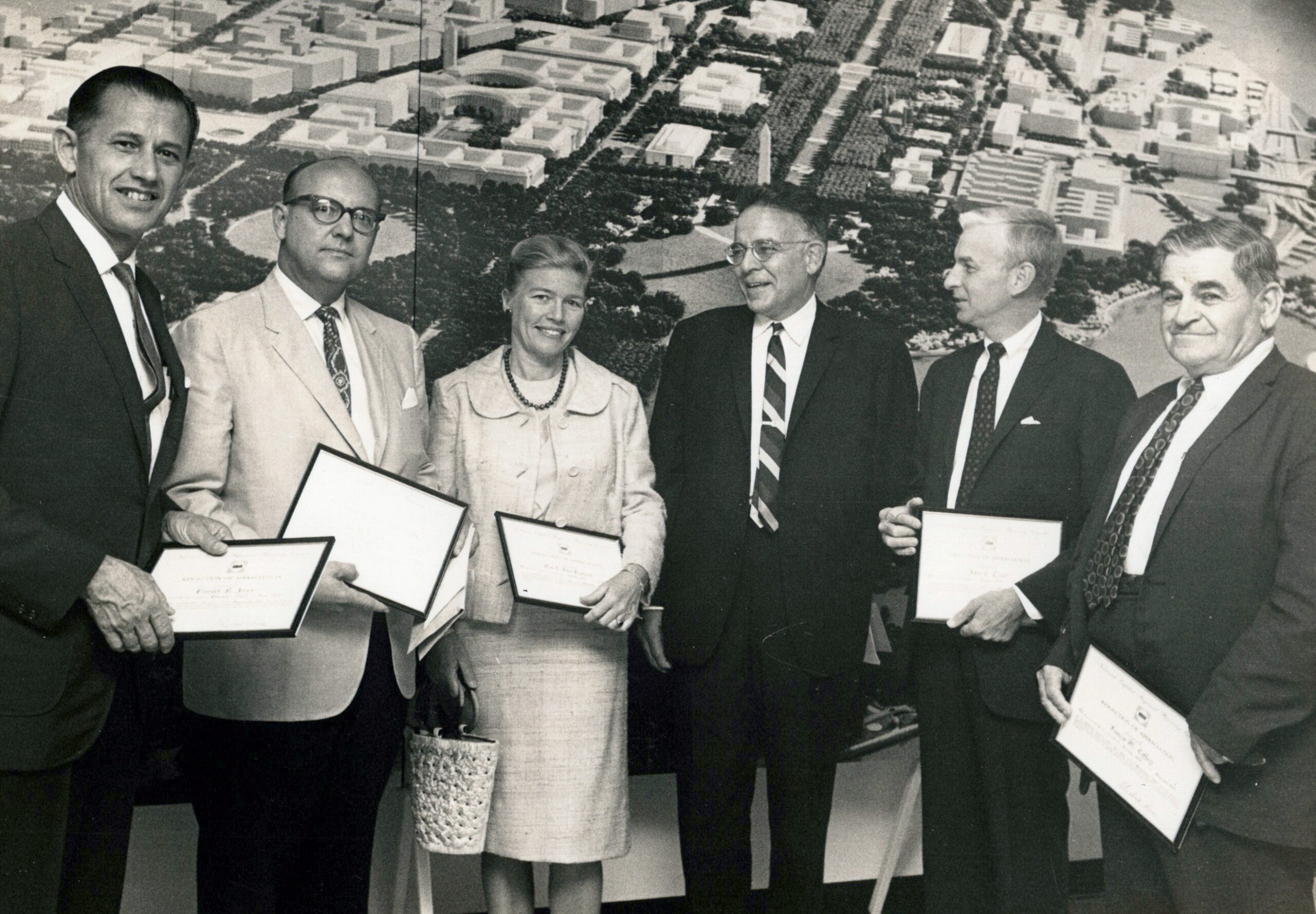 A black-and-white photo of Caroline Freeland standing with male colleagues. Most hold a framed certificate. They stand in front of an aerial photo of Washington, DC.