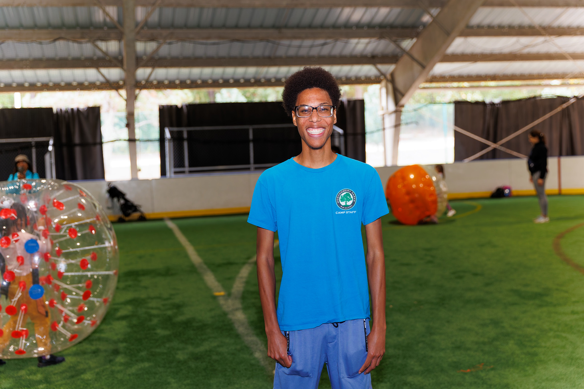 A person is standing on a sports field with children playing with bubble bump balls in the background.