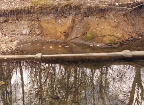 An exposed sewer line is partially submerged in a stream. A heavily eroded stream bank in background.