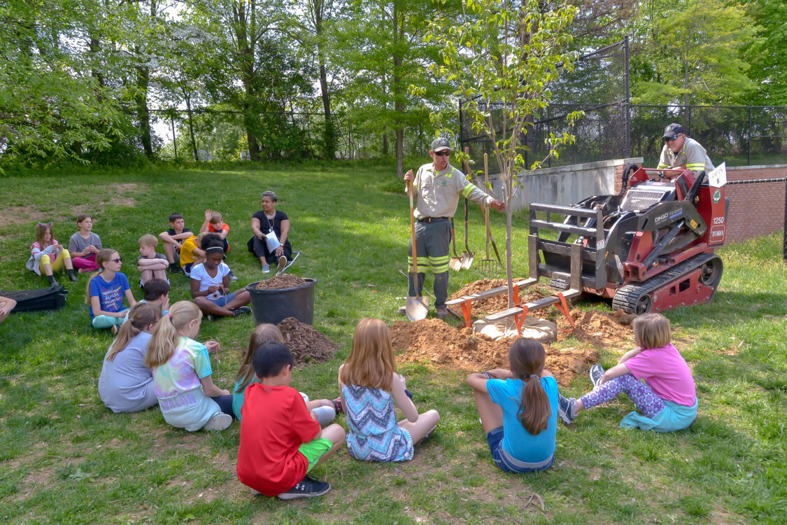 Children sitting around two arborists displaying some tools they use