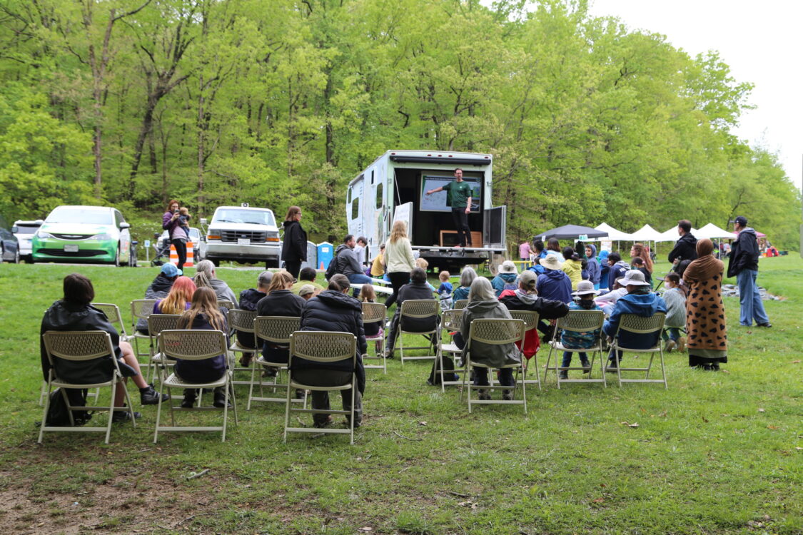 A scientist presents a talk about park ecosystems to an outdoor audience from the Nature on Wheels stage.