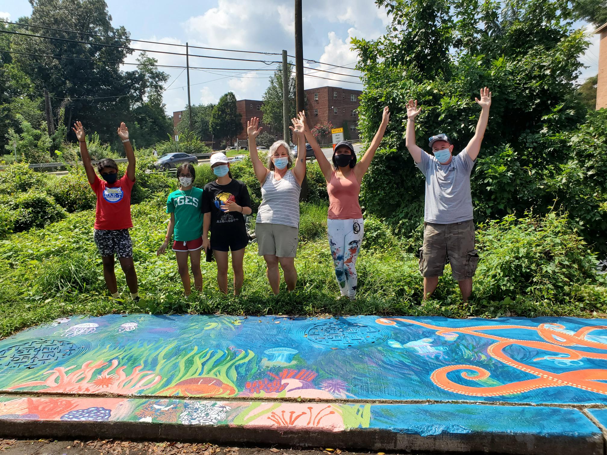 volunteers at a park raising hands and standing over a storm drain mural. the mural has images of the ocean featuring an octopus, ruby, holding litter items at each arm.