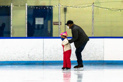 Adult helps child skate on indoor ice rink.