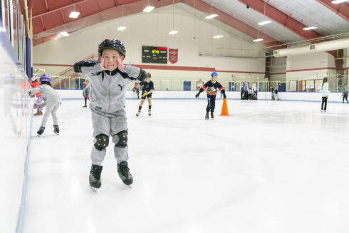 Child stops ice skating at indoor ice rink and looks into camera smiling with hands placed on both cheeks.