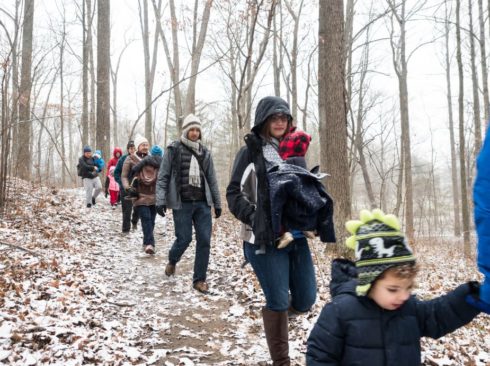 Adults and children bundled up walking through a snowy wood on a natural surface trail.