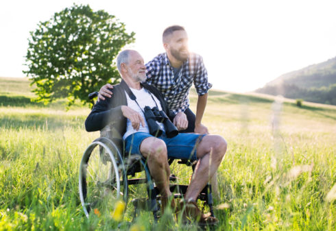 An adult leans over and has his arm around an older adult in a wheelchair. The older adult has binoculars around his neck. They are outdoors in a meadow, and both look into the distance while smiling. 