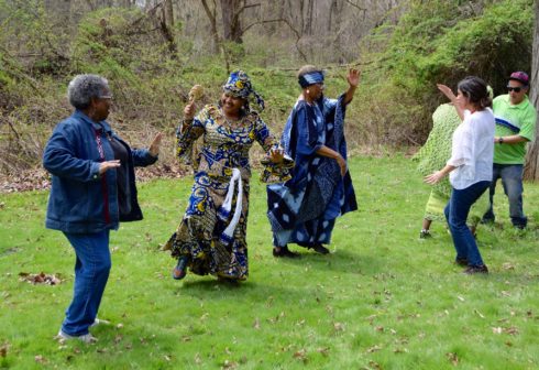 Four people dancing. Two people in the middle are dressed in outfits made from African prints. Each dancer faces one other dancer. They are all outdoors.