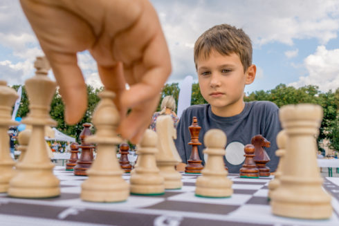 A hand close to the camera hovers above a bishop chess piece. A boy across the board focuses on other chess pieces.