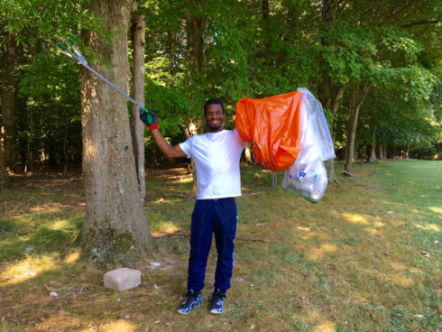 A young adult stands holding two garbage bags in one hand and a grabber for litter in the other. He is picking up trash in a park.