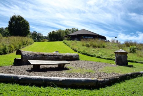 A bench, log, and small wooden house which make up a child's nature play area at the bottom of a grassy hill from Black Hill Visitor Center.
