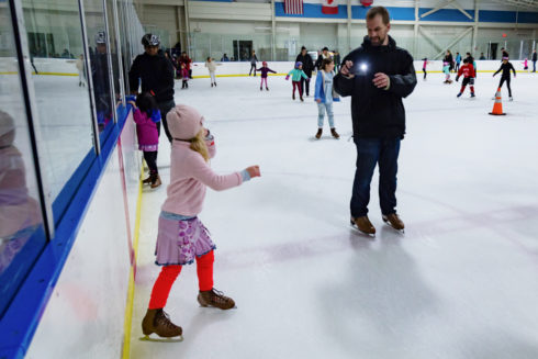 An adult and child at a public ice skating session. The adult takes a photo of the child with his phone. 