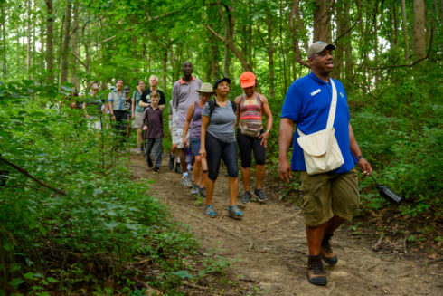 A staff member in a blue shirt with a satchel over his should leads a group of adults and children on a hike through the woods.