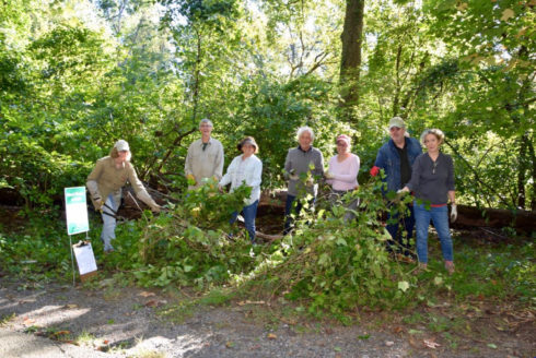 Seven adults hold vines and stand in front of piles of vines and other plants removed as part of a Weed Warrior workday.
