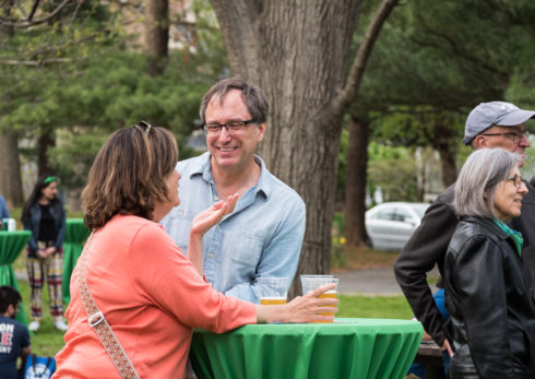 Two adults smiling and talking over cups of beer at an outdoor beer garden in a park.