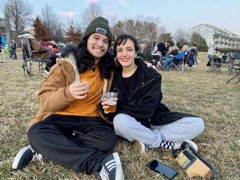 Two young adults smile and hold beers in plastic cups while sitting cross-legged on the ground.