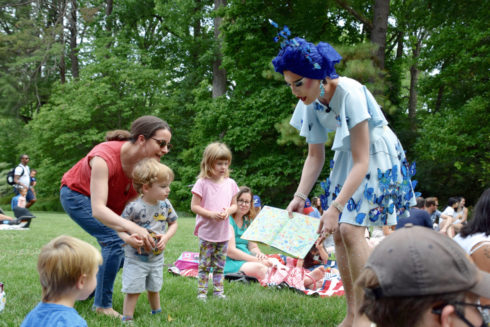 A drag queen in a blue dress with butterflies and blue head wrap bends over to show an open book to an adult and two children. They are outdoors and others are sitting on the lawn around them.