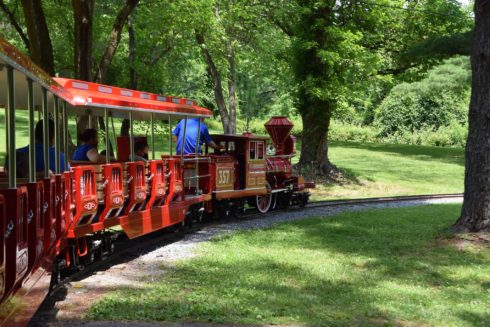 A red engine pulls red cars filled with passengers through Wheaton Regional Park.