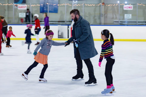 A parent holds the hand of a child who is skating backwards. Another child looks on.
