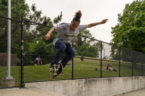 A teen jumps into the air on his skateboard at a skate park. There are people sitting on a hill in the background watching skaters.
