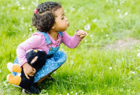 Small child squats down in a grassy area. She holds a dandelion and blows the puffball of seeds.