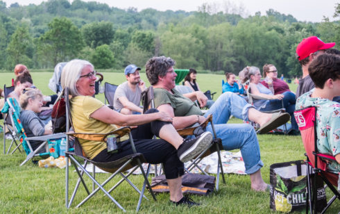 People sitting in camping chairs outdoors. They are laughing while attending a comedy show in a park.