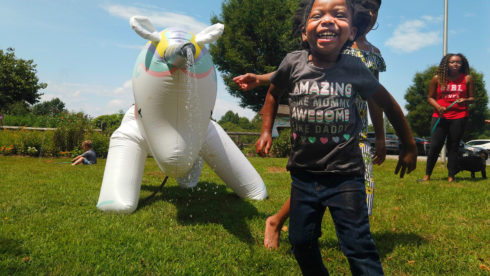 Smiling toddler runs away from a blow-up unicorn sprinkler. Sprays of water come out of its horn pointed towards the child.
