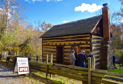 A small group of people is getting a tour from a guide in front of the historic Oakley Cabin. There is a sign in front of the home which says "Oakley Cabin. Open."
