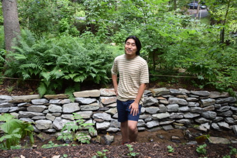 Summer intern Sanghyuk stands outdoors near a stream. Behind him is a stone retaining wall and many bushes and ferns. 