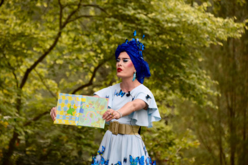 Drag queen stands outdoors in a blue dress and head wrap adorned with butterflies. She holds up an open book and shows to audience.
