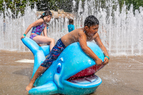 A child climbs on a whale water feature at South Germantown SplashPark and MiniGolf. Two other children play in water sprays in the background.