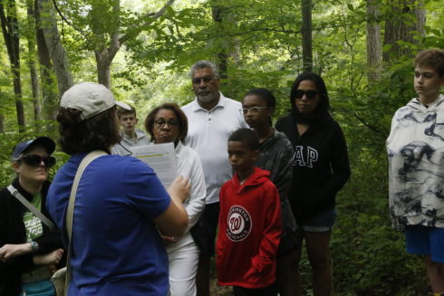 Adults and children watch and listen to a guide on the Underground Railroad Trail Guided Hike. They are outdoors in the woods.