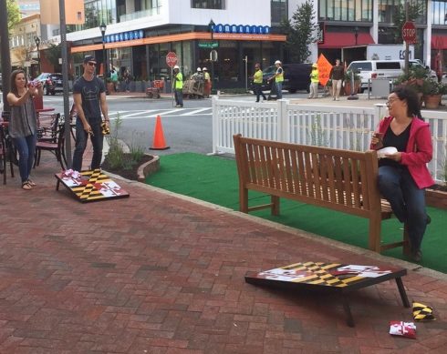 Two people play cornhole while another watches seated on a bench.