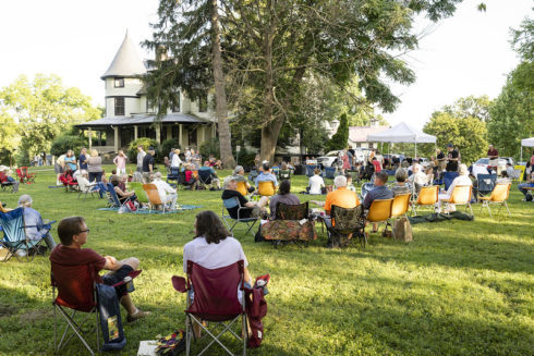 People sitting in camp chairs listening to an outdoor concert in a park. There is a Victorian nineteenth century house with a turret in the distance.
