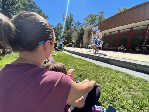 A child sits in the lap of an adult listening to a drag queen read a story. The reader holds the book high for the audience to see. The program is outdoors on a sunny day.