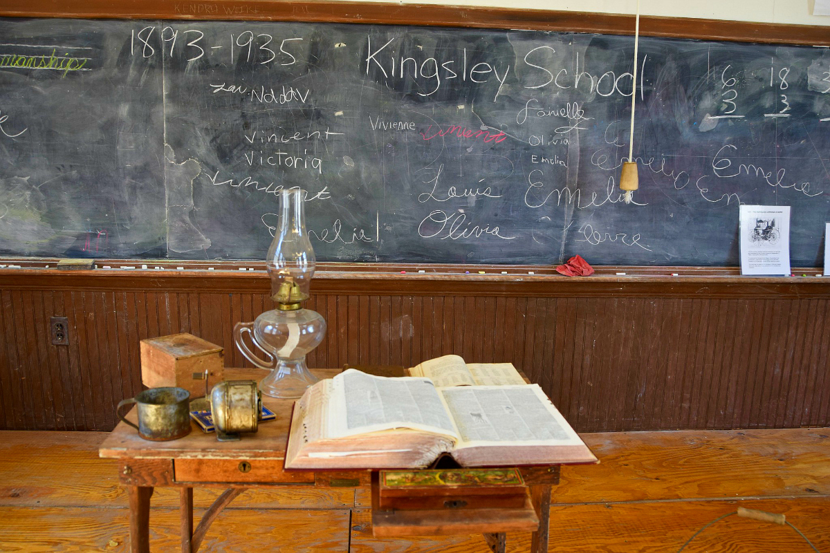 A large blackboard with writing inside a late nineteenth century one-room schoolhouse. In the foreground is a small desk with an open book, kerosene lamp, and other items.