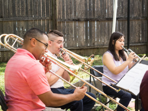Two trombone players and a trumpet player perform outdoors in a park.