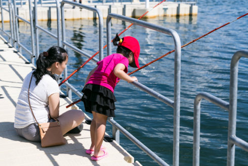 A parent sits on a boat ramp while a child looks into the water. They both have fishing poles. 