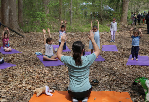 Adult and children on yoga mats in the woods. They all have their arms above their heads pointing skywards.