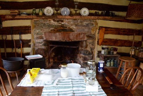 Stone hearth inside the historic Oakley Cabin. A wooden table has a white table cloth over it, and it is set with items for baking or cooking. 