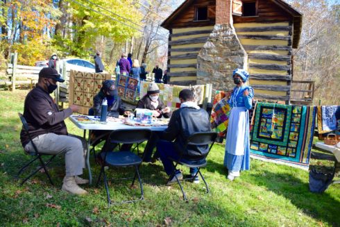 Adults and children sit at a table making quilt squares for a quilt project. Quilts hang on a line behind them. There is a woman in period dress talking to the family. And, behind all of them is the historic Oakley Cabin.