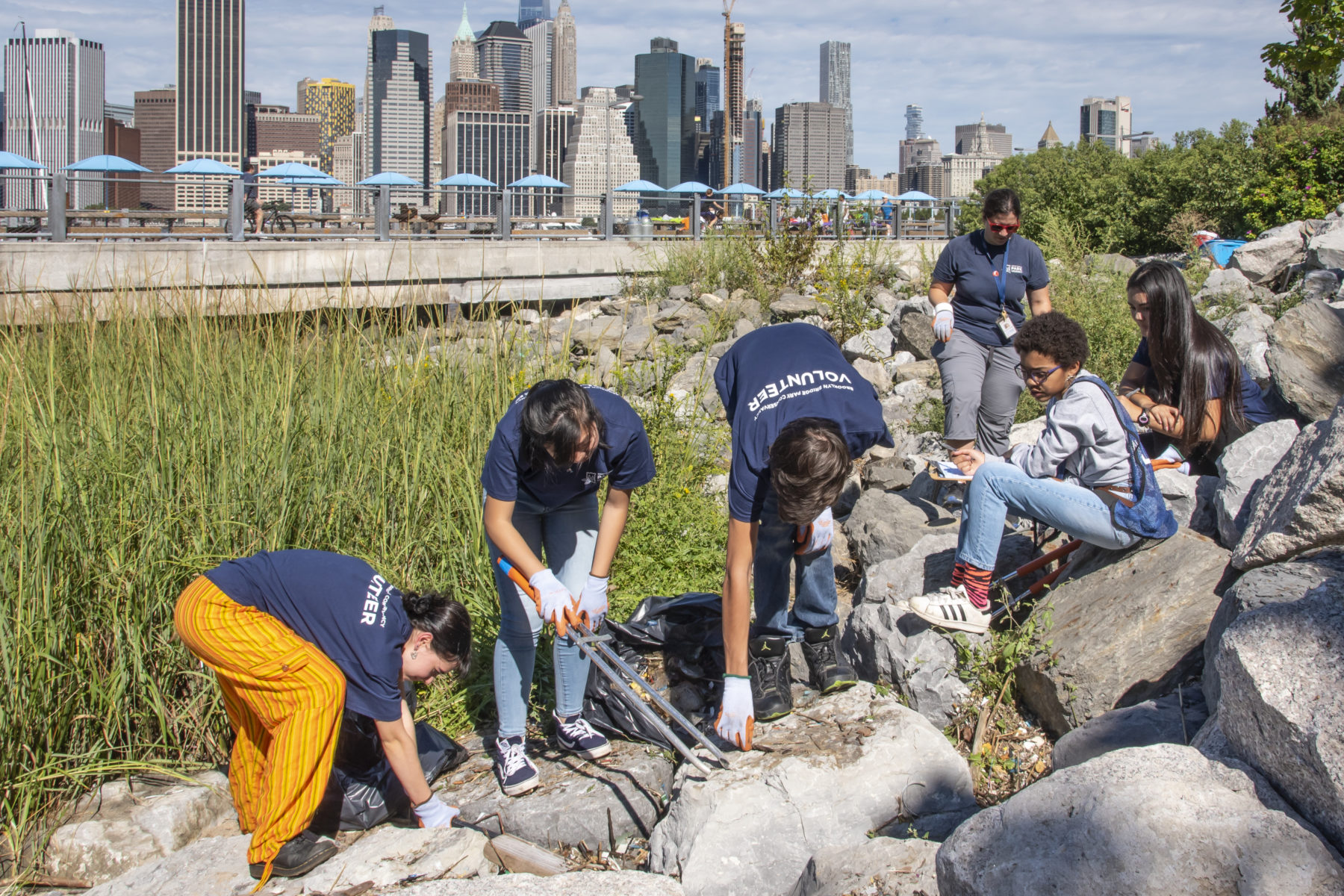 Teenagers cleaning trash from rocks in a park against the New York City skyline.  