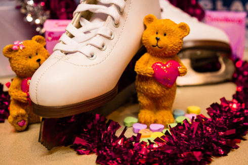 A pair of figure skates on a table. They are flanked by ceramic bears holding red hearts. Tinsel hearts surround them, and candy hearts are sprinkled near one.