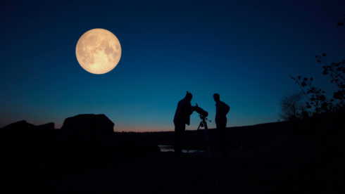 Two people stand and look at a full moon through a telescope. There are the outlines of two buildings in the distance.