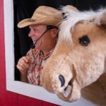 Kidsinger Jim with his head out of his barnyard trailer window accompanied by his Henry the horse puppet