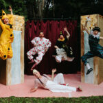 preposterous cast members posing in front of their set pieces in costume (4 members leaping in the air and one member lying on his side on the ground underneath them)