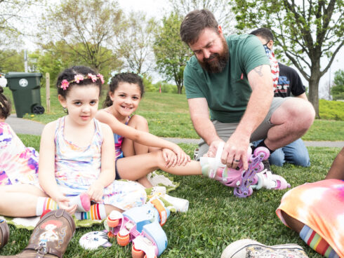 Dad helping daughters strap on their rollerskates as they sit on a grassy hill posing for a picture