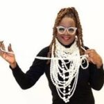 miss chocolate posing in a black turtleneck dress adorned with dozens of strings of pearls, and wearing white glasses