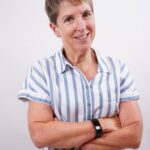 shot of sandi bersntein with arms folded smiling at the camera in a white and blue striped button down short sleeve shirt