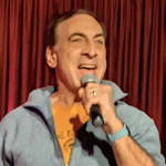 photo of walter performing on stage with a mic in his hand, red curtain in the background, and a gray quarter zip with yellow t shirt underneath