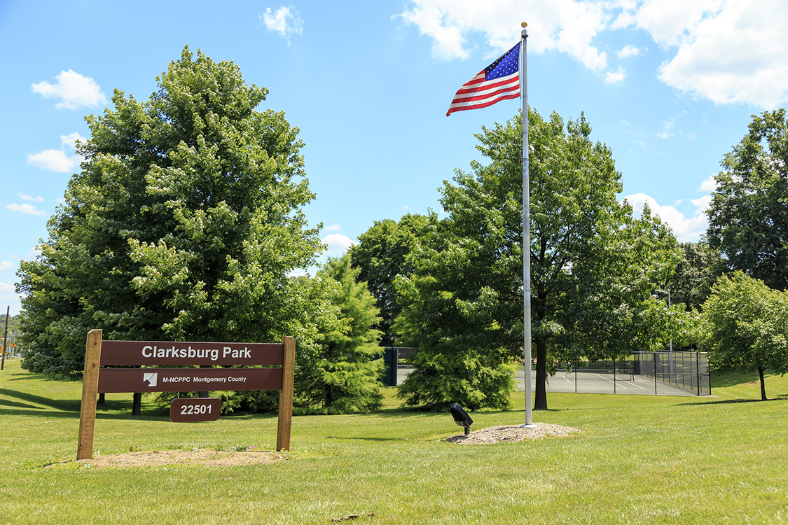 The brown Clarksburg Park sign next to the flying flag and tennis courts.